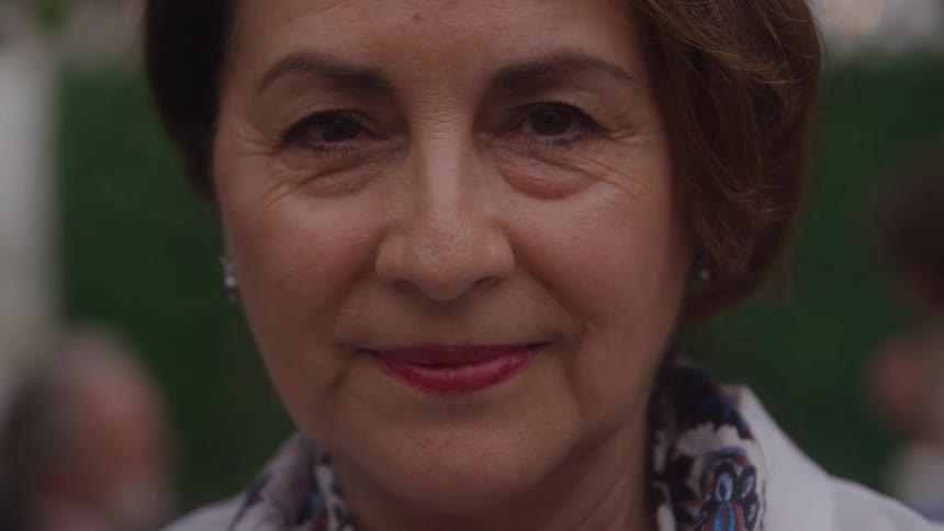 A closeup shot of a womans face with short brown hair and a patterned scarf shows her looking directly at the camera with a warm closedmouth smile against a softly blurred green background with undistinguished people