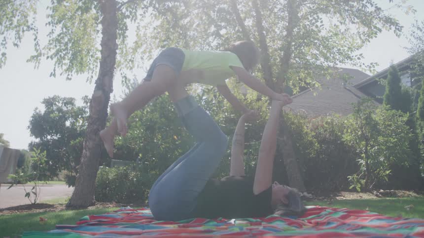 A woman wearing a dark shirt and jeans lies on her back on a colorful blanket and lifts a young girl wearing a yellow shirt and shorts over her using her hands and feet on a sunny suburban lawn with trees and houses visible
