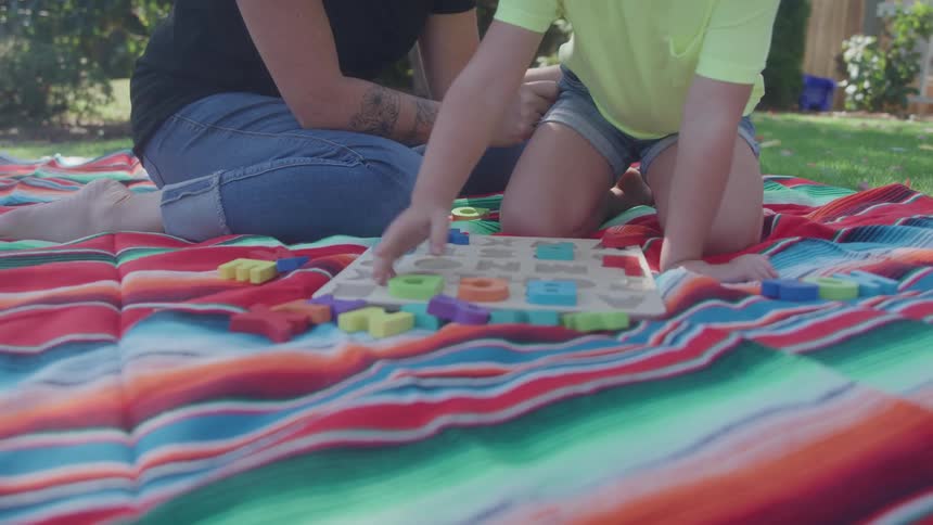 Child engages with educational toy on striped blanket beside adult in grassy area
