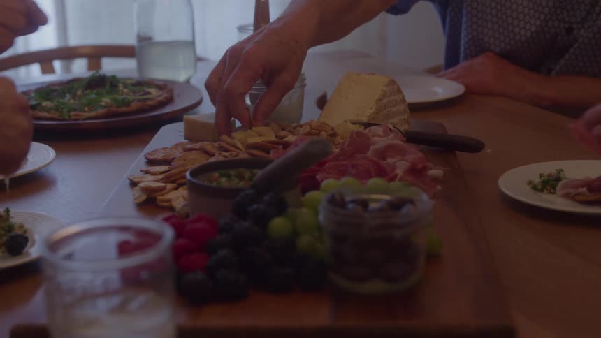 A charcuterie board laden with cheese crackers cured meat and fresh berries and grapes sits at the center of a wooden dining table as three peoples hands reach in to take pieces of food and talk during a bright daytime meal