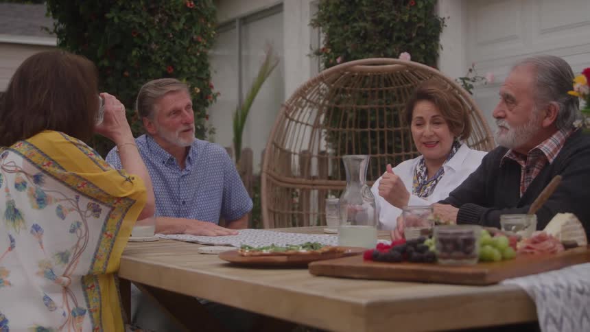 Group of elderly people talks around a table with food in an outdoor area surrounded by greenery