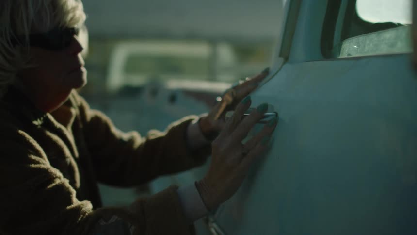 A woman with dark glasses feels along the surface of a truck near the door handle