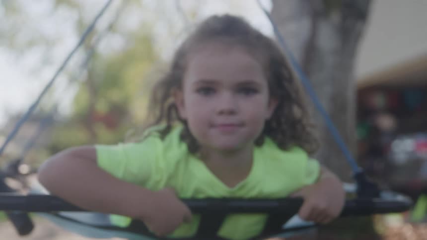 Little girl in green shirt holds edges of suspended swing in outdoor area with tree in the background