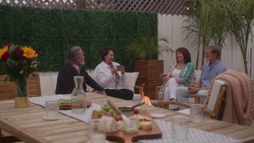 Four adults sit in chairs and on a couch around an outdoor fire pit and side table engaging in conversation with a dining table in the foreground a white wooden fence and green artificial hedge