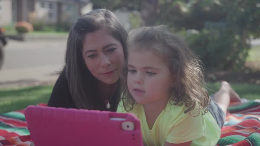 A mother with dark hair and a young girl with curly hair lie close together on a colorful blanket on a sunny suburban lawn looking intently at a pink digital tablet that the child is operating