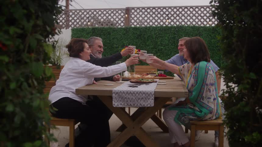 Four adults sit at an outdoor wooden table with drinks and food framed by green foliage in the foreground and raise their glasses to each other in a toast or cheer against a green hedge backdrop and pale sky