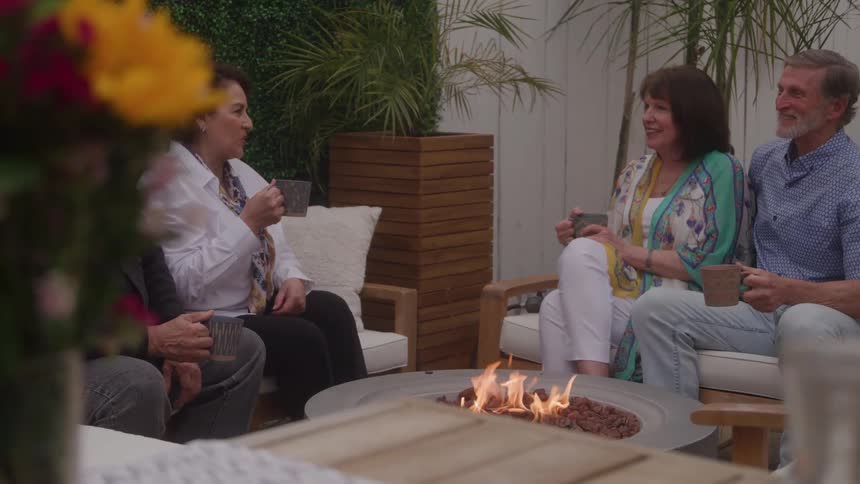People toast with mugs around a fire table in an outdoor area decorated with plants