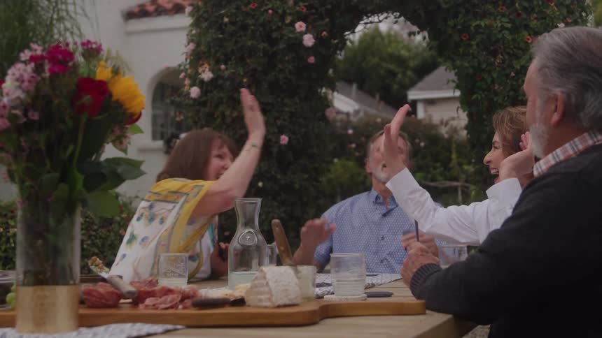 Two elderly men and two middleaged women sit at an outdoor table under the midday sun laughing as the women highfive after winning a lively card game