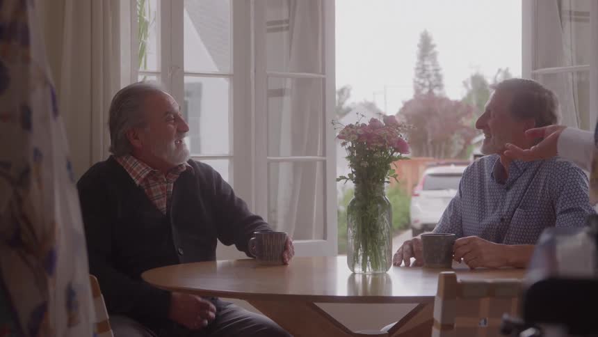 The video clip features two older men sitting at a round wooden table having a casual and cheerful conversation they are both holding mugs and smiling suggesting a warm social gathering or a pleasant chat in a bright sunlit room with a vase of flowers