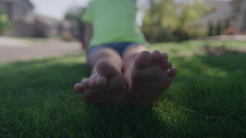 A lowangle video shows the bare feet of a young child in a lime green shirt and shorts sliding into focus and resting on a patch of lush green grass in a sunny residential lawn with blurry houses visible in the distance