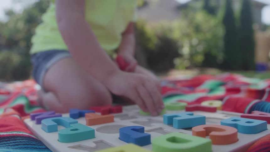 Child kneeling on colorful blanket fits blue letter into alphabet puzzle in backyard with greenery
