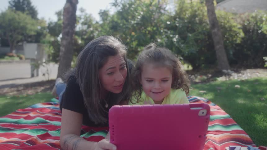 A mother and her young daughter with curly hair lie on their stomachs on a colorful striped blanket on a sunny green lawn smiling while looking closely at a pinkcased tablet in this outdoor family video