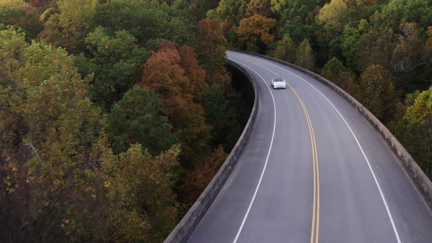 An aerial view video shows a white car driving slowly around a long curving overpass highway surrounded by a dense forest canopy beginning to show autumn colors of green gold and red