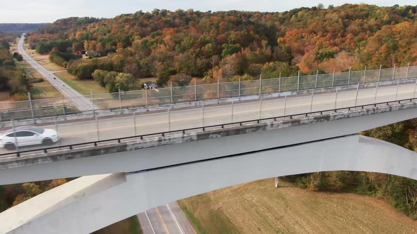 The video is an aerial wide shot showing a modern arch bridge crossing a highway and a valley landscape the surrounding hills are covered with autumn foliage featuring a mix of green yellow and red trees