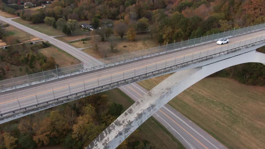 White car crosses an elevated bridge over a highway surrounded by trees