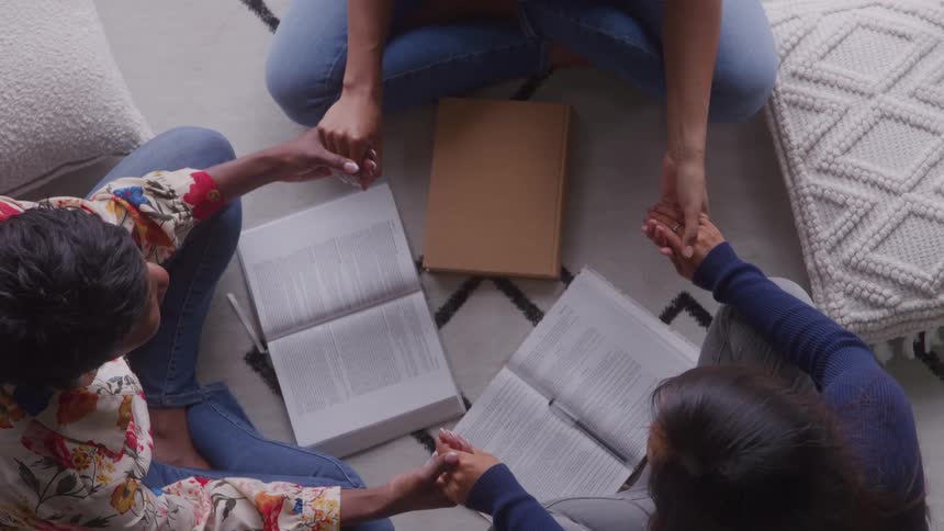 Three people sitting in a circle holding hands surrounded by open books