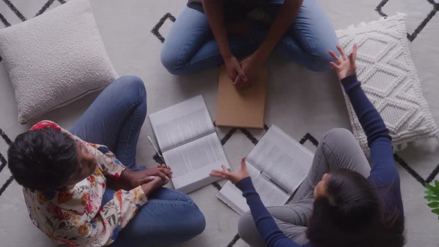 An overhead view video shows three women in jeans sitting crosslegged on a white patterned rug leaning over and reading large textbooks together while studying with white decorative pillows scattered nearby in a bright room