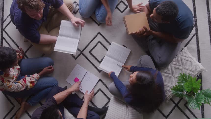 Five young adult friends sit in a circle on the floor indoors engaged in a relaxed book reading session sharing passages and thoughts in a cozy inviting atmosphere