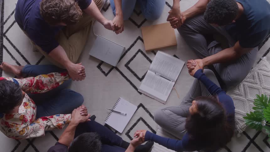 An overhead shot shows six people sitting on a patterned rug in a circle with hands joined with books and notebooks in the center as the group appears to be praying
