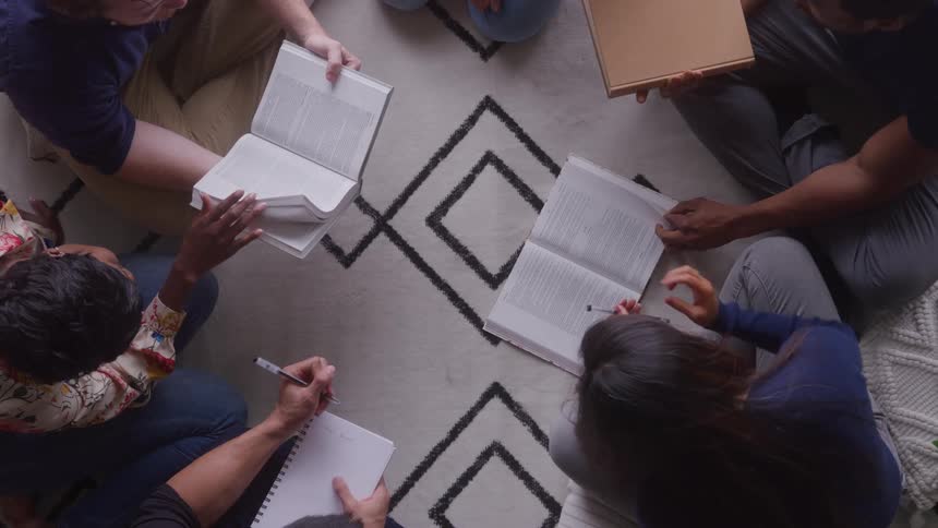 The video is an overhead shot of five students sitting in a circle on a patterned rug actively studying or working on a project they are surrounded by books notebooks and papers emphasizing a collaborative learning environment