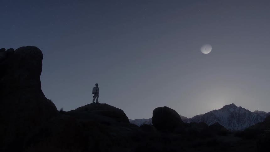 The silhouette of a person walking on a large dark rock looking at the sky on a clear morning with a large moon