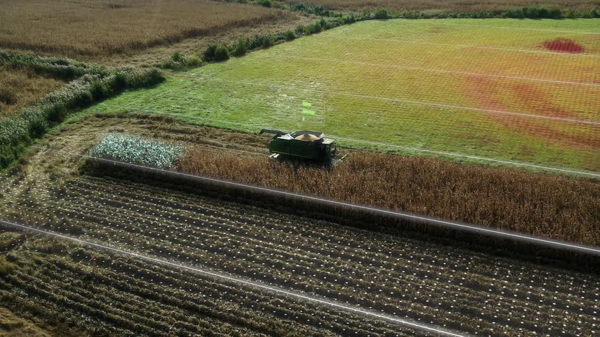 An overhead drone video shows a combine harvester with a bin full of grain moving through rows of dry brown crops with a visual overlay of white digital lines and green data points indicating precision agriculture in the surrounding fields