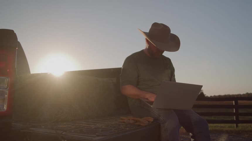 Man wearing a cowboy hat uses a laptop while sitting on a truck bed at sunset