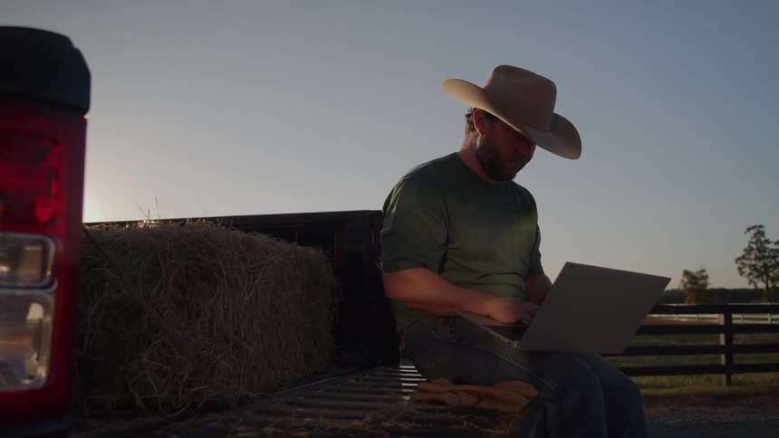 Man wearing a cowboy hat uses a laptop while sitting on a truck bed