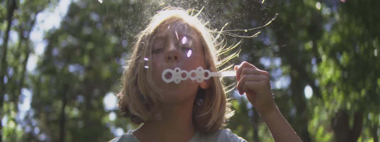 A young boy stands in a woodland at midday using a bubble wand to shimmering bubbles into the sunlit air surrounded by trees and playful light