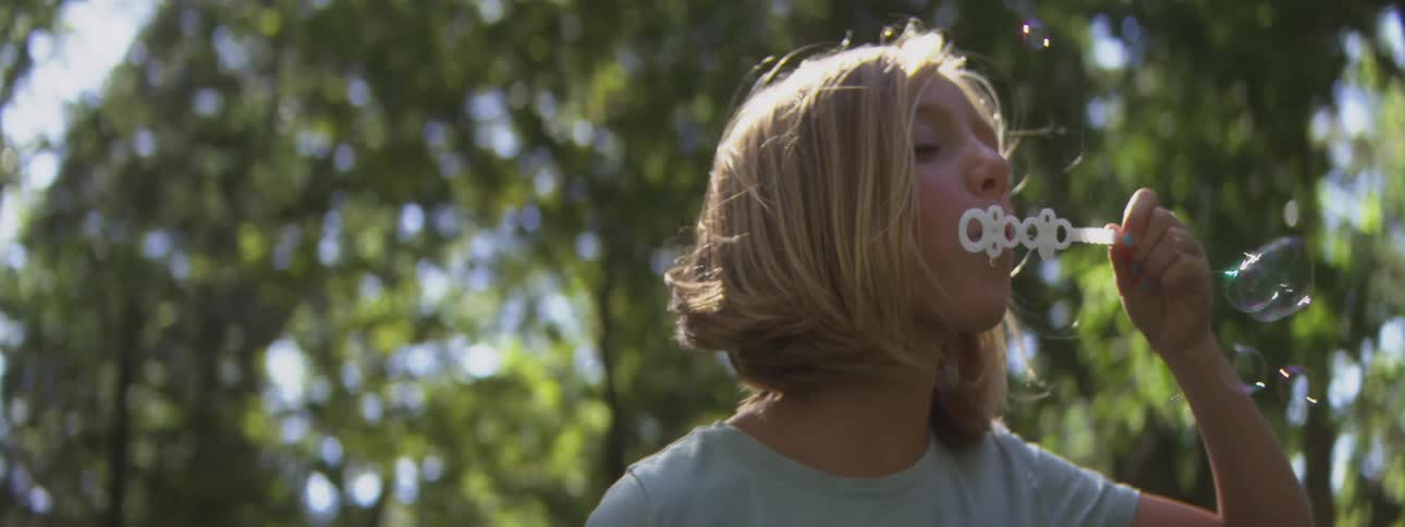 Child blowing soap bubbles outdoors with blurred trees in the background