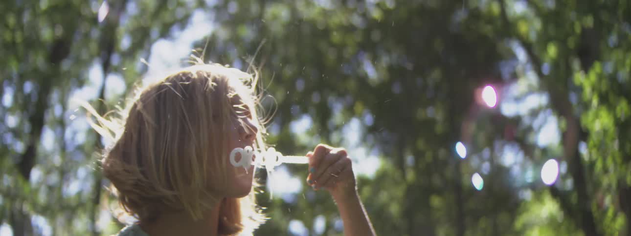 A young boy holds a bubble wand apparatus to mouth and exhales creating multiple soap spheres that float into the air while standing among tall plants