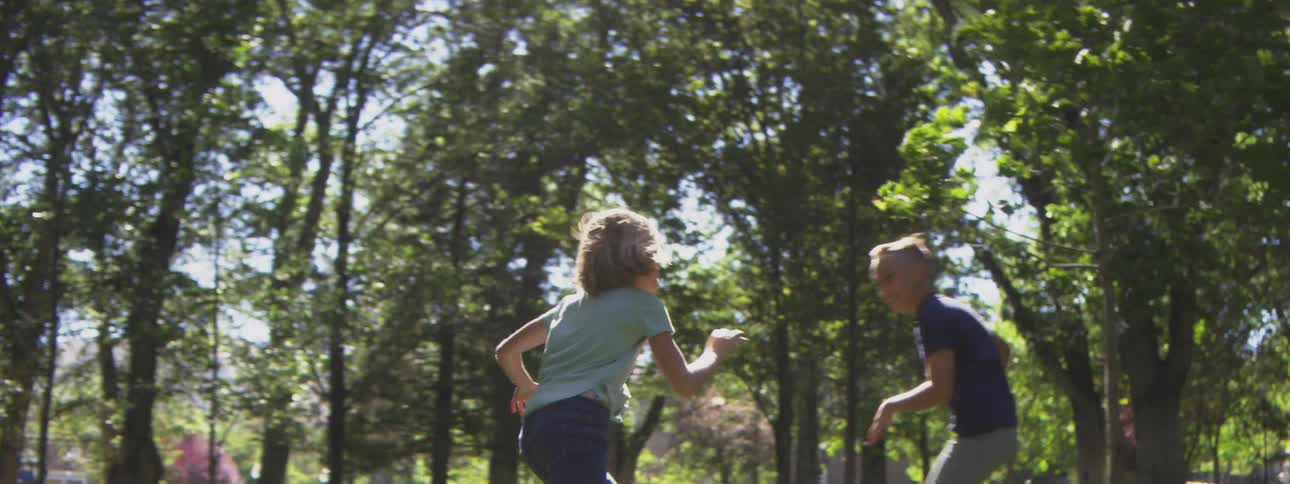 Two children a boy in a dark shirt and a child in a light shirt run and jump on a patch of grass surrounded by trees while playing a chasing game in a public park setting during the day