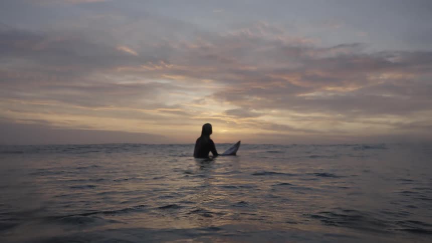 A person sits on a board in the water with their back to the camera positioned on the ocean surface below a sky with stratified cloud formations near the time when the sun is below the horizon