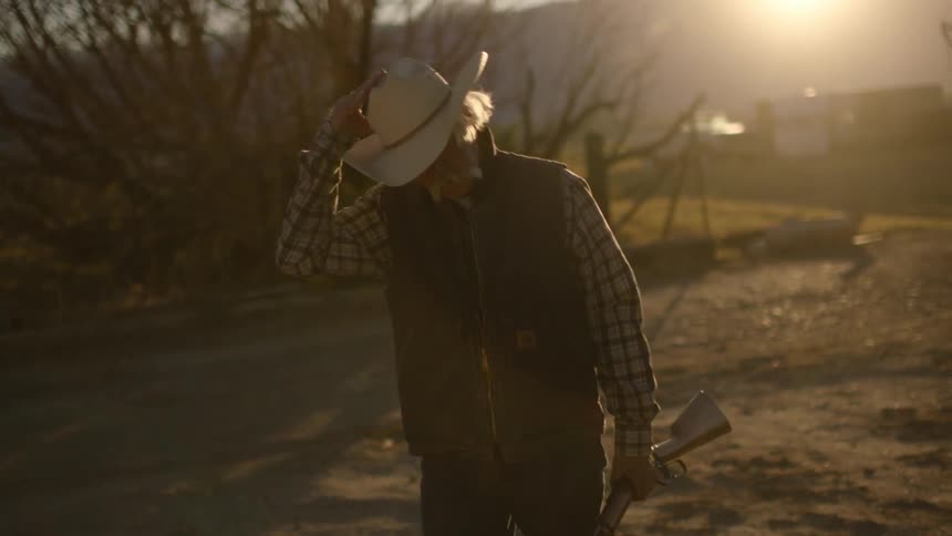 Elderly man holding a gun walks over his property at sunset