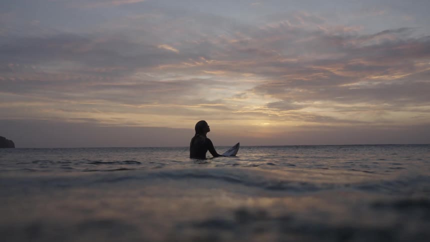 Woman sitting on her surfboard in the water looks at the sky at dawn