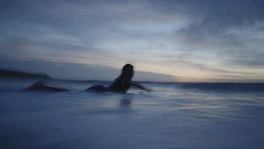 The video is a lowangle shot of a person paddling a surfboard or boogie board out in the ocean at dusk or dawn the water is foamy and dark and the low light casts the surfers figure in silhouette against the pale sky