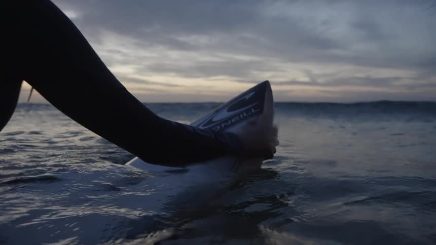 The video shows a surfer in a black wetsuit at the oceans surface holding the nose of their surfboard the scene is set during twilight or sunset with the person silhouetted against the dark sky and calm water