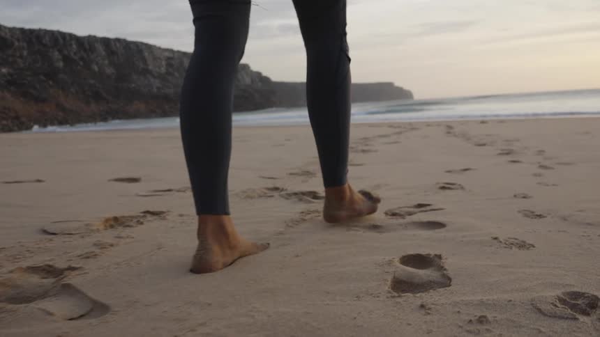 A person in a wetsuit walks barefoot with a surfboard on a wide sandy beach fresh footprints trailing toward the shoreline with rugged cliffs in the distance
