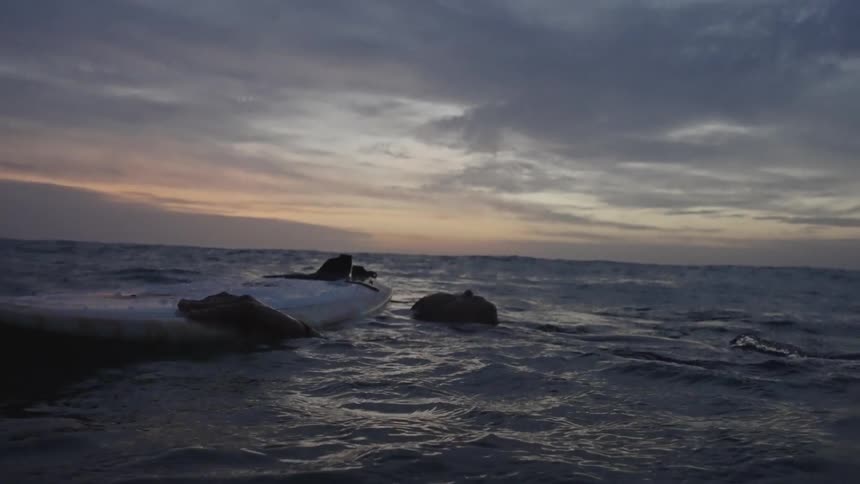 A pair of surfers in wetsuits float calmly on their boards at dusk beneath a wide pastel sky over gently rippling water