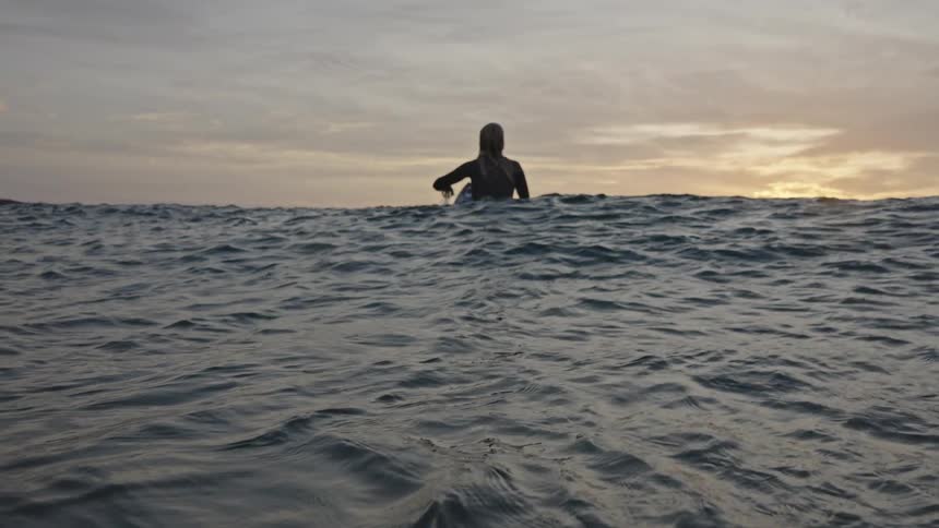 A woman sits on a board in the water facing away from the camera on the surface of the moving ocean at the sunset