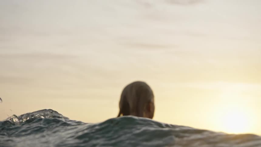 A woman wearing a dark fullbody garment lies flat on a board in the water and uses arms to propel the board forward on the ocean surface during the time of day when the sun is near the horizon