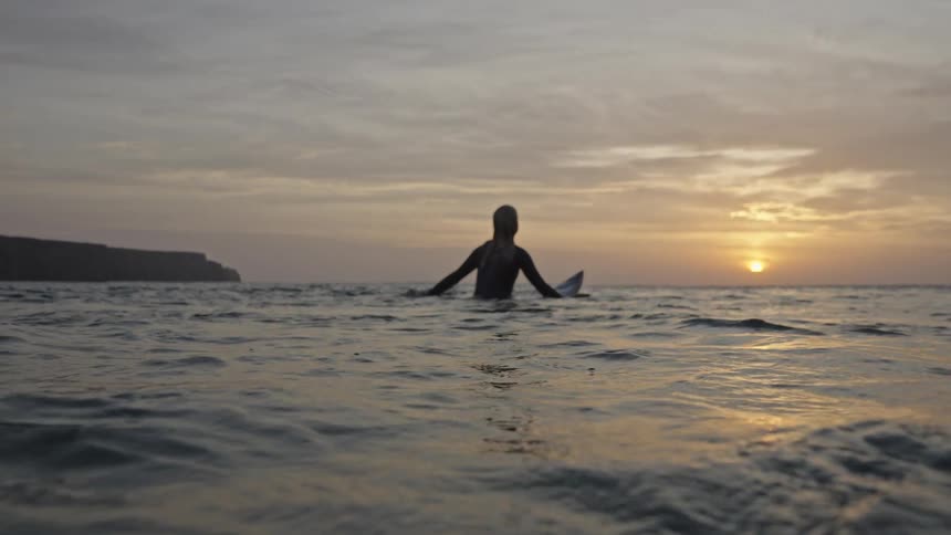 The video shows a surfer sitting on their board in the ocean silhouetted against a sunset or sunrise the horizon features a dark prominent landmass or cliff on the left and the bright sun low on the right creating a peaceful moody sce