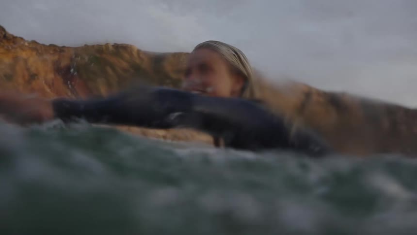A woman in a wetsuit paddles on a surfboard by rocky coastal cliffs smiling with relaxed joy as small waves ripple around her