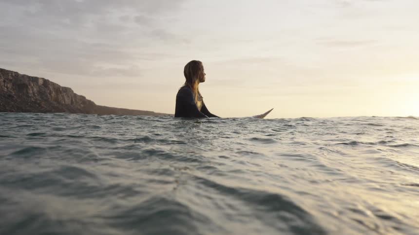 A woman in a wetsuit sits astride a surfboard in calm golden water near rugged cliffs gazing toward the bright horizon
