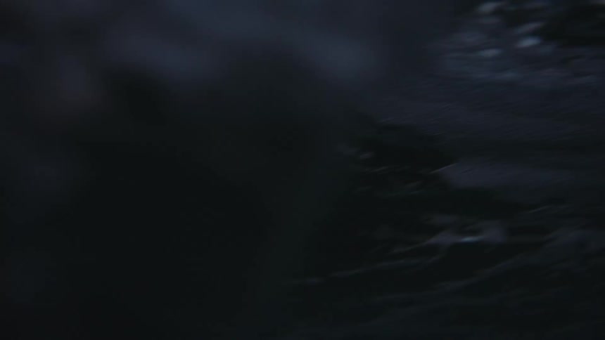 The video is a lowangle shot that captures a wave breaking in the ocean at twilight the sky above is a mix of dark gray and pale light and the rocky shoreline is visible in the distance to the left
