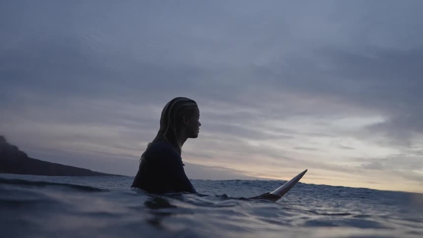 The video shows a person sitting on a surfboard in the ocean at dusk or dawn silhouetted against a cloudy colorful sky the lowangle perspective captures the gentle movement of the dark water around them and the looming presence of a rocky shor