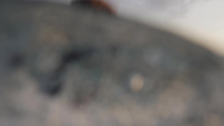 The video is a lowangle closeup shot of a person with blonde hair surfing or boogie boarding in the ocean they are paddling into a wave with water splashing around them and a dramatic rocky cliff face is visible in the background against a cl