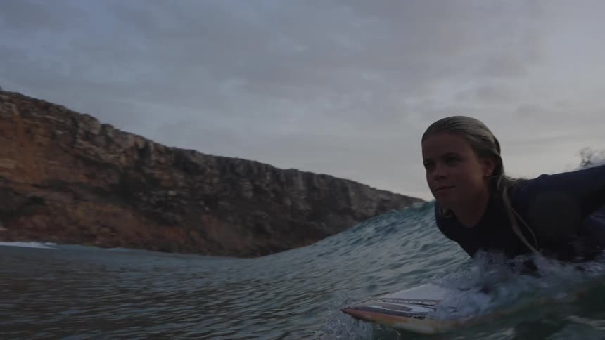 A water level close view shows a blonde woman wearing a dark wetsuit paddling a surfboard before catching and riding a wave toward a brown cliff face on an overcast day