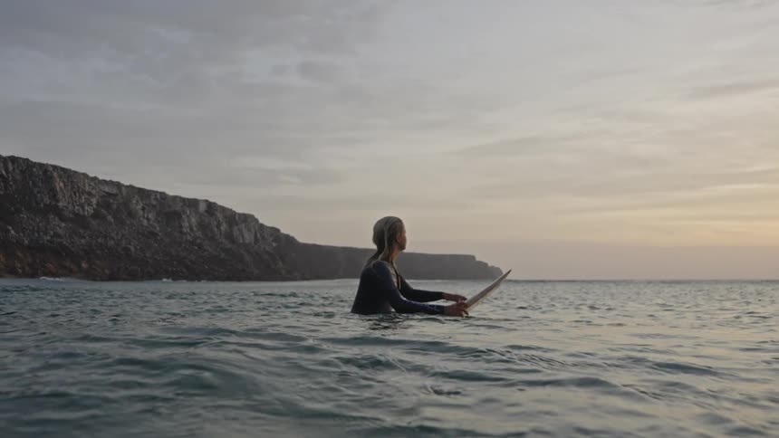 A young woman wearing a dark wetsuit sits on a surfboard in the ocean water with the waterline in the foreground facing the open sea against a cloudy fading sky and a large dark cliff in the background