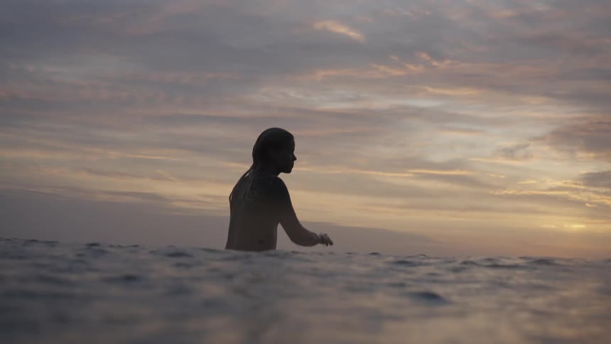 The video shows a silhouetted person submerged chestdeep in the ocean at either sunrise or sunset the sky is a hazy blend of yellow orange and purple clouds with the figure appearing to speak into a hand held up to their ear